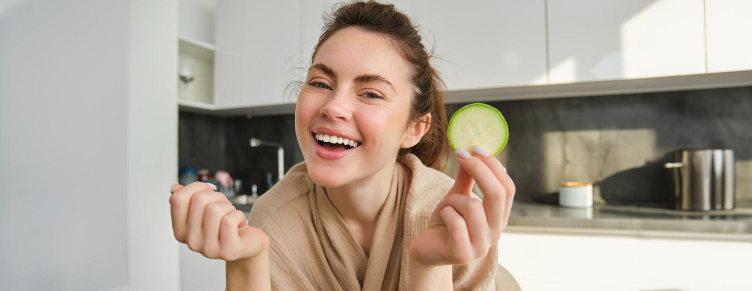 Eine lachende Frau in einem Bademantel hält in einer modernen Küche eine Gurkenscheibe in der Hand.