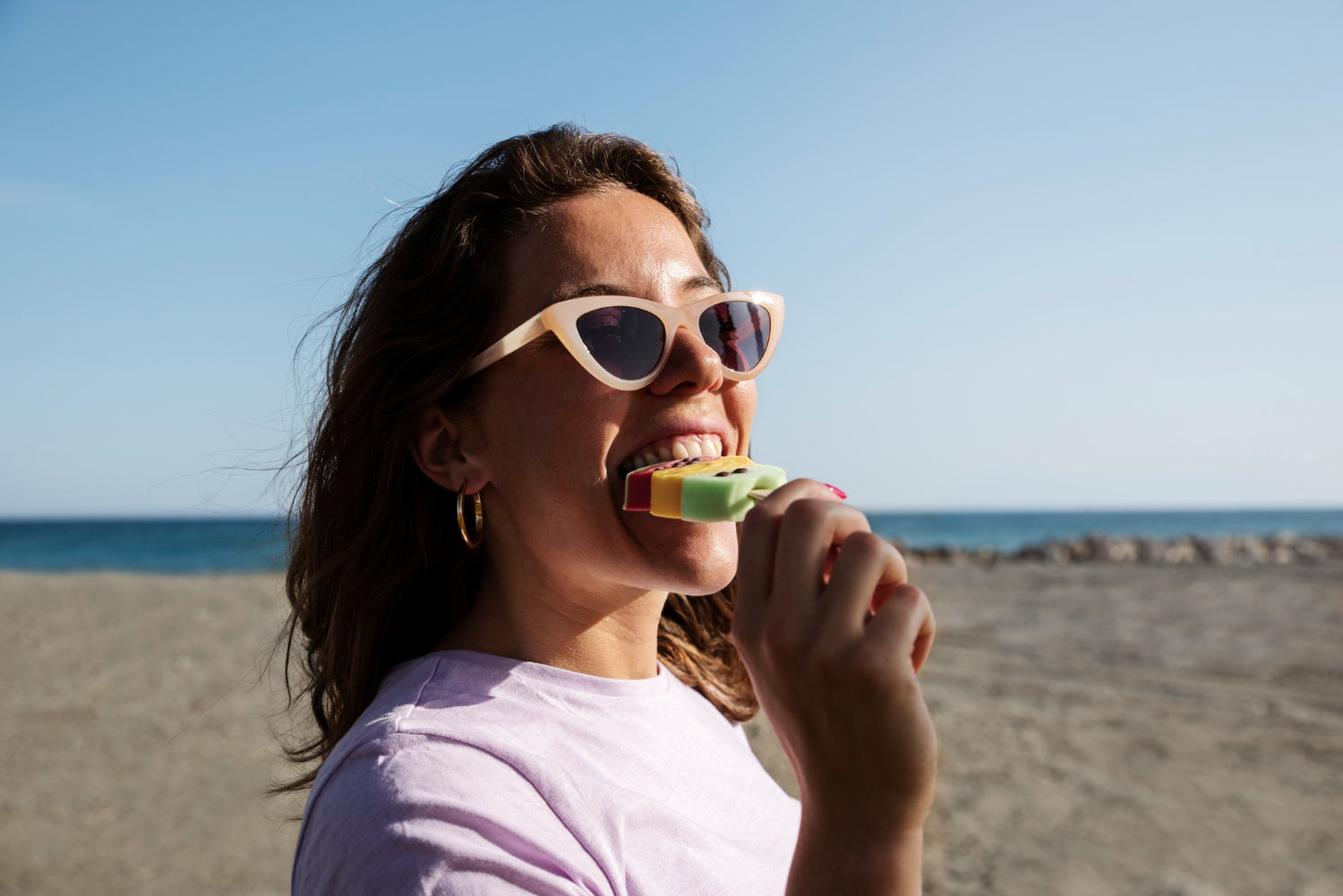 Eine Frau isst am Strand ein Eis und trägt Sonnenbrille und Creolen.