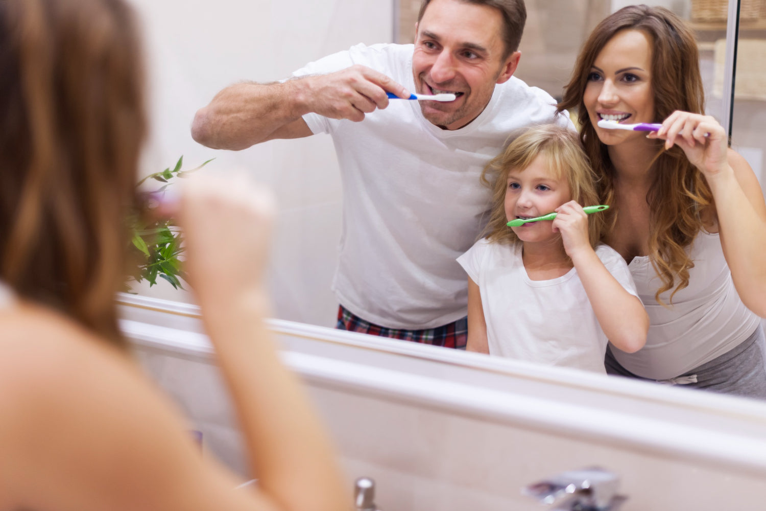 Eine Familie steht gemeinsam vor dem Spiegel im Badezimmer und putzt sich lachend die Zähne.