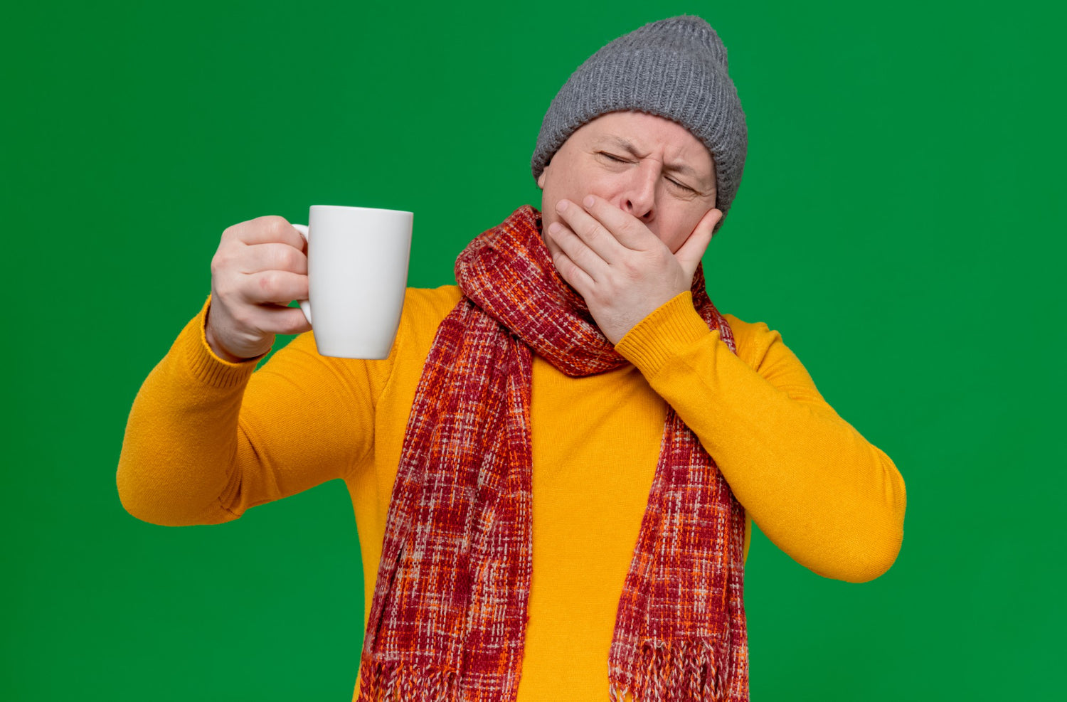 Eine Person mit Mütze und Schal hält eine Tasse in der Hand und gähnt vor einem grünen Hintergrund.
